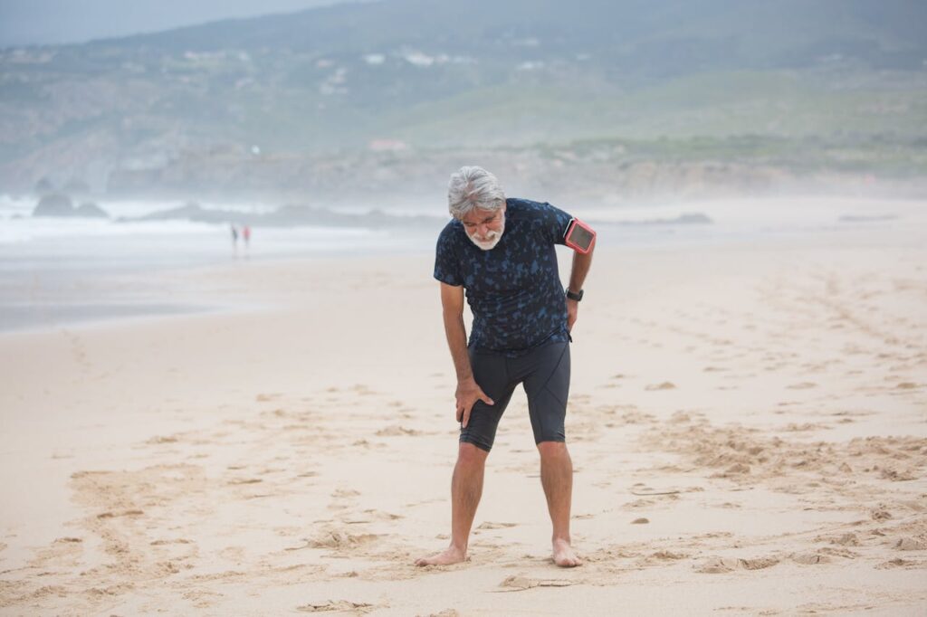 Older man leaning forward on a foggy beach, hand on lower back and thigh in visible sciatica pain, representing common lower back and leg nerve issues and how chiropractic provides natural, non-invasive relief at ChiroCure Health Center in Jacksonville, FL